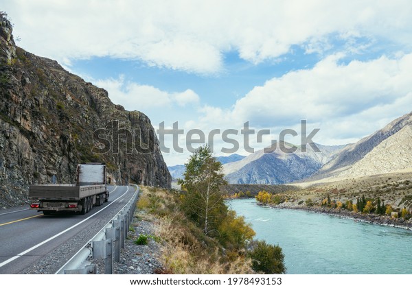Truck on mountain highway along big mountain river
in sunshine in autumn. Wide turquoise river and mountain road.
Highway in mountains. Russia, Altai Republic, Chuysky tract, 19
September, 2020.