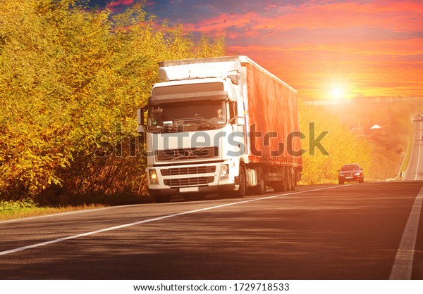 Truck\
with a red trailer and a car on an autumn countryside road with\
trees and bushes against a night sky with a\
sunset