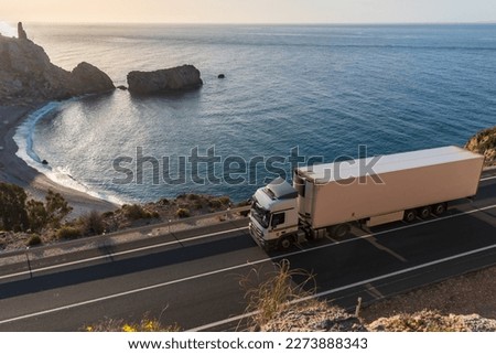 Truck with refrigerated semi-trailer driving along a road by the sea with a beach in the background.