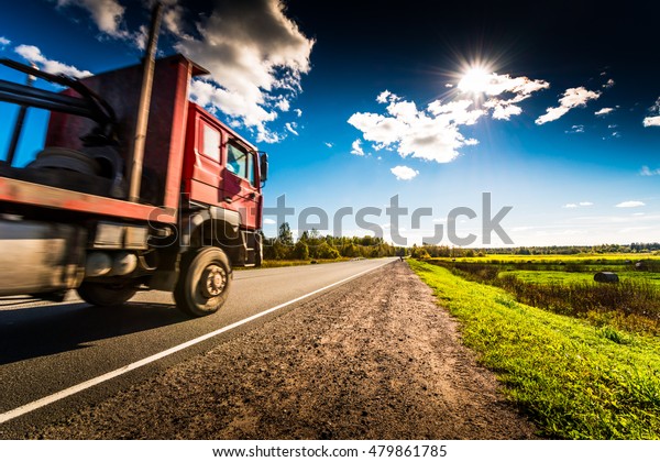 Truck rides along the forest road which is in the\
field with haystacks lit by the sun on a blue sky background. High\
contrast and obscured\
image