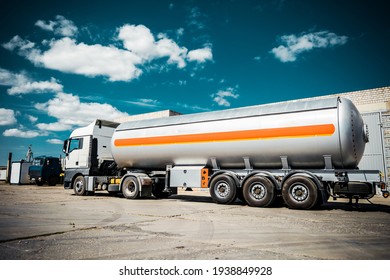 Truck with trailer, tank with flammable liquid, sunny day outside, metallic color container, blue sky with white clouds, gravel	