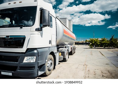 Truck with trailer, tank with flammable liquid, sunny day outside, metallic color container, blue sky with white clouds, gravel	