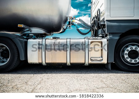 Truck with trailer, tank with flammable liquid, sunny day outside, metallic color container, blue sky with white clouds, gravel