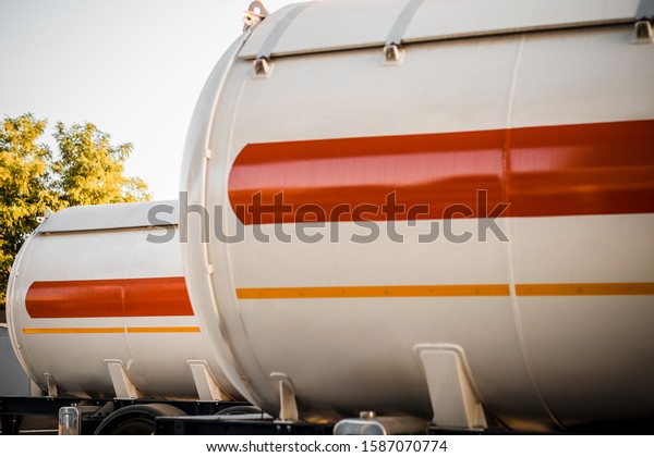 Truck with trailer, tank with\
flammable liquid, under a large bridge at the pier on the river\
bank, sunset light, white and red cars standing on\
gravel