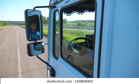 Trucker In Sunglasses Reflecting In Side Mirror Of Moving Car. Man Driving Lorry Riding To Destination Through Countryside Road. Cargo Transportation Concept. Beautiful Background. Slow Mo Close Up