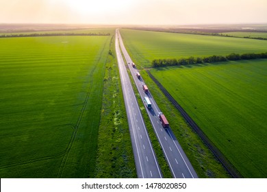 Trucks On The Higthway Sunset. Cargo Delivery Driving On Asphalt Road Along The Green Fields With Goods. Seen From The Air. Aerial View Landscape. Drone Photography. Convoys With Cargo