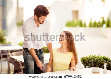 True gentleman! Young handsome brunet lover is adjusting the chair of his happy lady, both well dressed, at a sunny terrace of restaurant