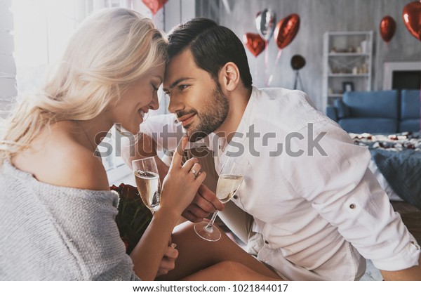 True love. Beautiful\
young couple drinking champagne and smiling while sitting face to\
face in the bedroom
