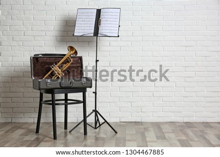 Trumpet, chair, case and note stand with music sheets near brick wall. Space for text