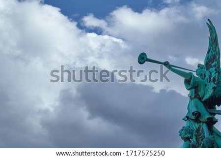 Trumpeting old bronze angel statue covered with green verdigris playing a celestial fanfare against a sunny sky with dramatic clouds.