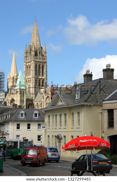 Truro Cornwall\
England. Street scene showing cars and local bus. Red cafe parasol.\
Tower of the cathedral in background rising over the roofs of\
nearby houses. Blue sky light\
cloud.