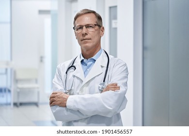 Trustworthy Proud Mature Old Medical Professional Doctor Stands Arm Crossed In Hospital. Middle Aged Male Senior Physician Practitioner Wears White Coat, Stethoscope, Glasses Looks At Camera. Portrait