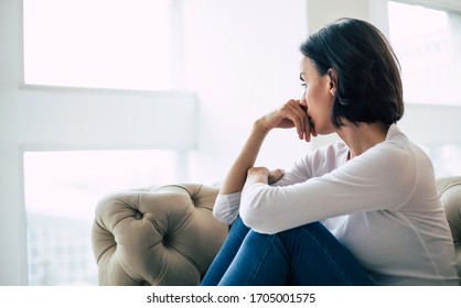 Trying to cope. Close-up photo of an adult woman who turned away from the camera, looking through the window with sadness. - Shutterstock ID 1705001575