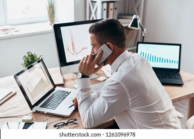 Trying To Help Someone. Young Businessman In Formalwear Talking On The Phone And Analyzing Data Using Computer While Sitting In The Office