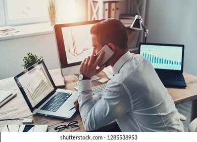 Trying To Help Someone. Young Businessman In Formalwear Talking On The Phone And Analyzing Data Using Computer While Sitting In The Office