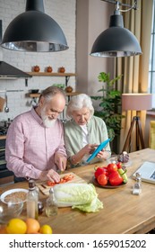 Trying New Meals. An Elderly Couple Using Recipes From The Internet While Cooking