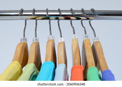 T-shirts of different colors hang on closing rack on wooden hangers with plastic size tags indexes of the XXS, XS, S, M, L, XL, XXL on a light background