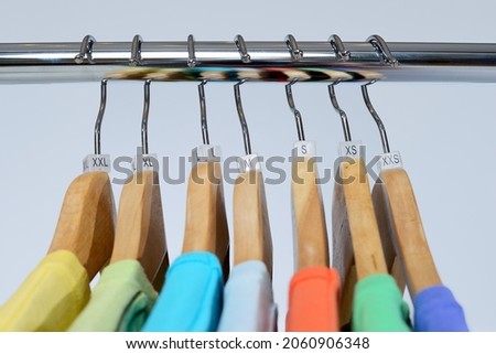 T-shirts of different colors hang on closing rack on wooden hangers with plastic size tags indexes of the XXS, XS, S, M, L, XL, XXL on a light background