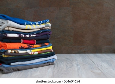 T-shirts are piled in a pile. A pile of T-shirts and sweatshirts is stacked on a wooden table. Clothes on a gray background. Clothes made of knitted fabric. T-shirts and sweatshirts are multicolored.