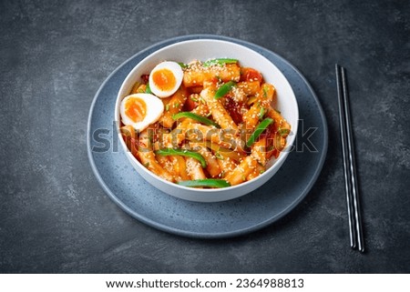 Tteokbokki with eggs, fresh green leek and roasted sesame seeds in blue bowl on the dark rustic table. Tteok-bokki is a Korean cuisine dish with rice gnocchi sticks and spacy sauce. Asian street food