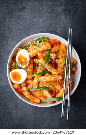 Tteokbokki with eggs, fresh green leek and roasted sesame seeds in blue bowl on the dark rustic table. Tteok-bokki is a Korean cuisine dish with rice gnocchi sticks and spacy sauce. Asian street food