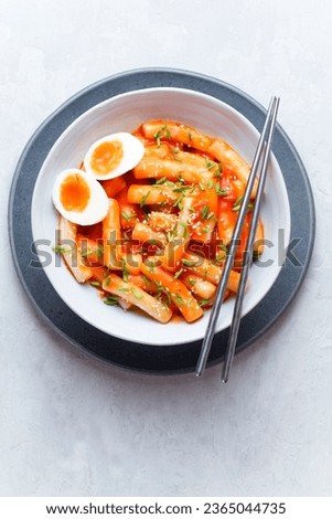 Tteokbokki with eggs, fresh green leek and roasted sesame seeds in blue bowl on the white stone table. Tteok-bokki is a Korean cuisine dish with rice gnocchi sticks and spacy sauce. Asian street food