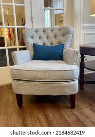 A Tufted Arm Chair That Is Grey With Steel Nail Heads Around The Border. It Sits In Front Of Glass French Doors And Has A Blue Lumbar Pillow On It.