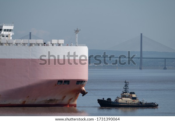A tugboat leads a giant car carrier ship across\
an estuary before a bridge.