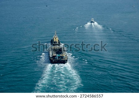 A tugboat pulls a cargo ship through calm waters close to the shore. The sky is clear, and the vessels create wakes as they move together. The scene shows activity on the water.