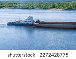 A tugboat pushes a large barge with cargo along a wide river. In summer, the tugboat goes down the river. Cargo transportation by river. Navigation of vessels on the river.