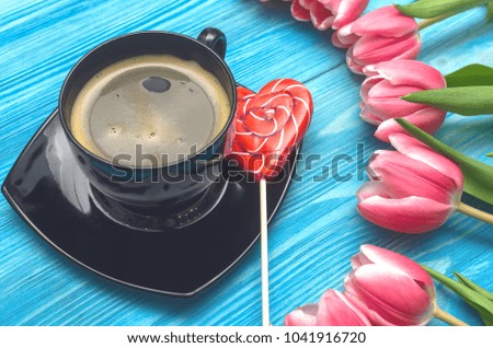 Tulip flowers and cup of hot black coffee with heart shape lollipop sweet on blue wooden background. Romantic breakfast concept background. Woman day. Saint valentines day backdrop top view.