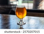 A tulip shaped clear beer glass filled with a cold blonde ale on a wooden table. The booze has a layer of froth on top of the liquor. The table is dark wood and there