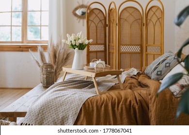 Tulips And Breakfast Tray On Bed With Teapot And Mug. Comfort Apartment In Bohemian Style Interior With Hygge Bedroom, Wooden Bench Seat, Bamboo Dressing Screen, Home Decor, Dry Plants In Vase