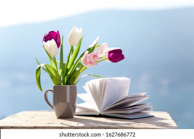 Tulips And Opened Book At Wooden Table Outdoors. Spring Still Life At Sea Beach. Blooming Flowers On Balcony At Blue Background Of Mountains. Pink, White, Purple Bouquet. Copy Space.