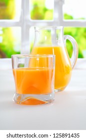 Tumbler Of Fresh Healthy Orange Juice Standing On A Kitchen Table Alongside A Jug Full Of Juice With A Sunny Window And Greenery Behind