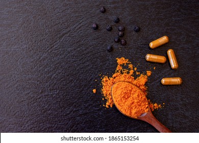 Tumeric powder in wooden spoon, Curcuma herbal capsule and black pepper isolated on dark table background. Health benefits and antioxidant food concept. Top view. Flat lay. 