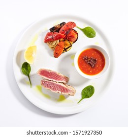 Tuna Steak With Grilled Vegetables Top View. Red Fish Piece With Tomato Sauce, Lemon Slices And Basil Leaves On Plate Isolated On White Background. Salmon With Cooked Veggies Composition 