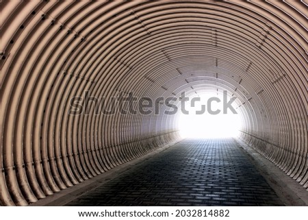 Tunnel in a hill made of metal blocks in the form of beige pipes, pedestrian pavement of concrete briquettes. The end of the tunnel is brightly lit by sunlight.