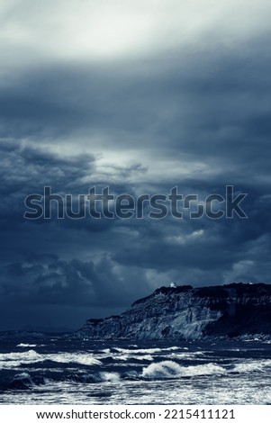 Turbulent ocean with a small house on a cliff under a dark cloudy sky.
