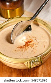 Turkey, Cauliflower And Apple Soup In A Glazed, Yellow Pottery Bowl With A Silver Spoon Dipped Into Liquid On A Wooden Cutting Board With A Pepper Grinder-Paleo Diet