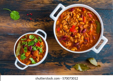 Turkey chili, ,stewed with beans, tomatoes, bell pepper, onion, garlic, thyme, cinnamon, chocolate and fresh cilantro, in white bowl and casserole on wooden table.