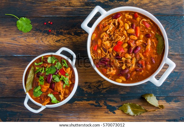Turkey chili, ,stewed with\
beans, tomatoes, bell pepper, onion, garlic, thyme, cinnamon,\
chocolate and fresh cilantro, in white bowl and casserole on wooden\
table.