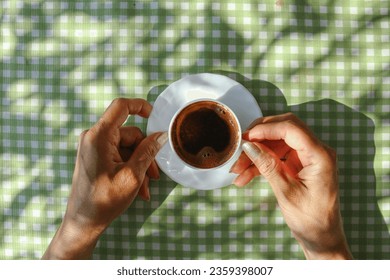 Turkish coffee in a white cup on a green and white square patterned tablecloth and a woman is holding this cup - Powered by Shutterstock