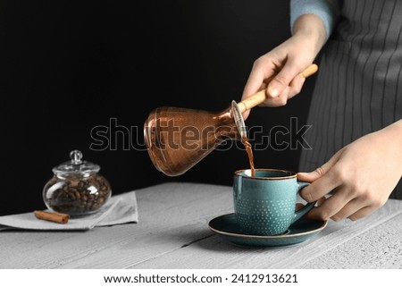 Turkish coffee. Woman pouring brewed beverage from cezve into cup at gray wooden table against black background, closeup