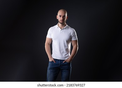 Turkish Guy With Bald Head Stand Focused With Neutral Face Expression Isolated On Dark Black Background With White Shirt While Look At Camera And Show His Deadpan Face  