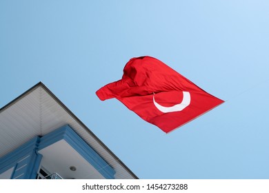 The Turkish Red Flag With The Moon And Star On A Background Of Blue Sky Surrounded By Buildings. Bottom View, The Flag Is Inverted, Fluttering In The Wind. The Concept Symbols Of The Countries.