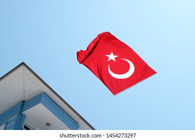 The Turkish Red Flag With The Moon And Star On A Background Of Blue Sky Surrounded By Buildings. Bottom View, The Flag Is Inverted, Fluttering In The Wind. The Concept Symbols Of The Countries.