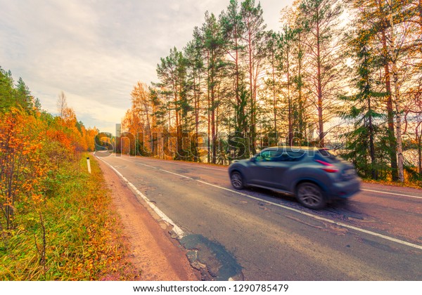 Turn the country broken road. The car goes on the\
road. Mixed forest. Sunset over forest lake. Autumn weather.\
Beautiful nature. Russia, Europe. View from the side of the road.\
Orange-purple toning.