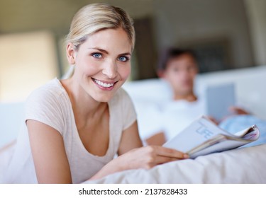 Turn The Page To A Happier You. Shot Of A Happy Young Woman Lying In Bed And Reading A Magazine While Her Husband Uses A Digital Tablet In The Background.