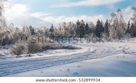 Turn of the snow-covered road among covered with fluffy snow trees. On the track visible tire treads of the wheels. In the blue sky white clouds. Photographed in winter in sunny weather. Soft focus